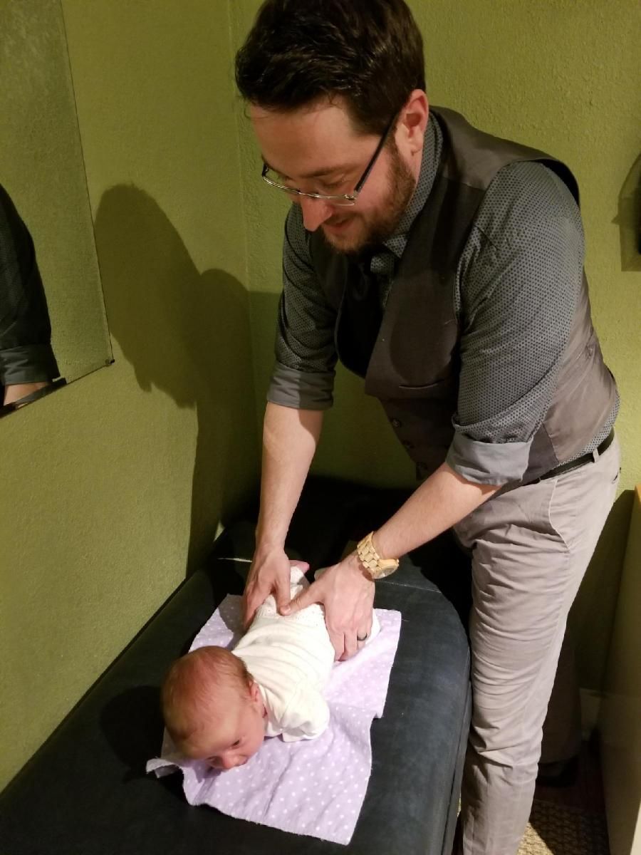 Man in glasses examining a baby on a table. He is dressed in light colors.