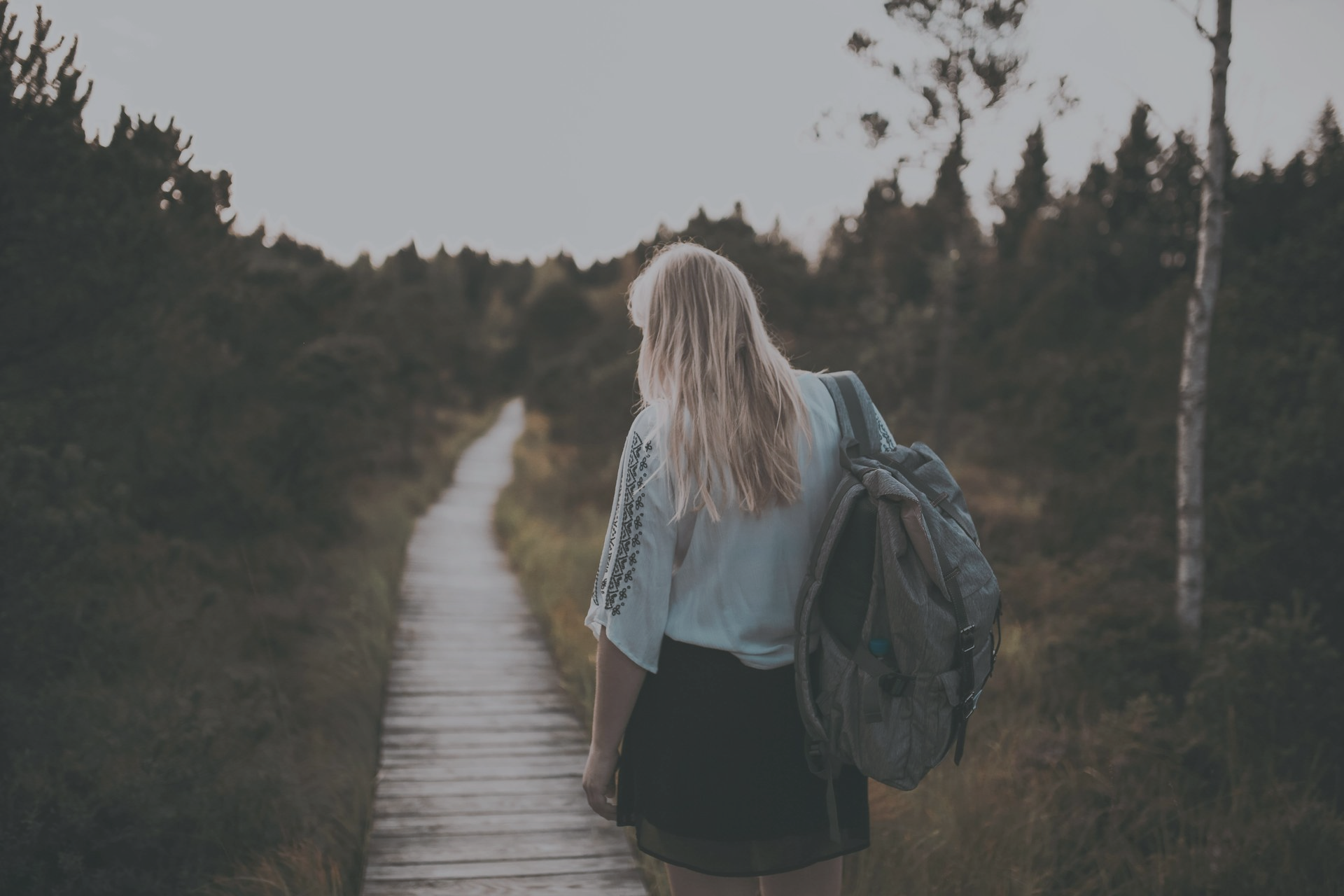Woman with blonde hair walks away down a wooden path, carrying a backpack through a forest.