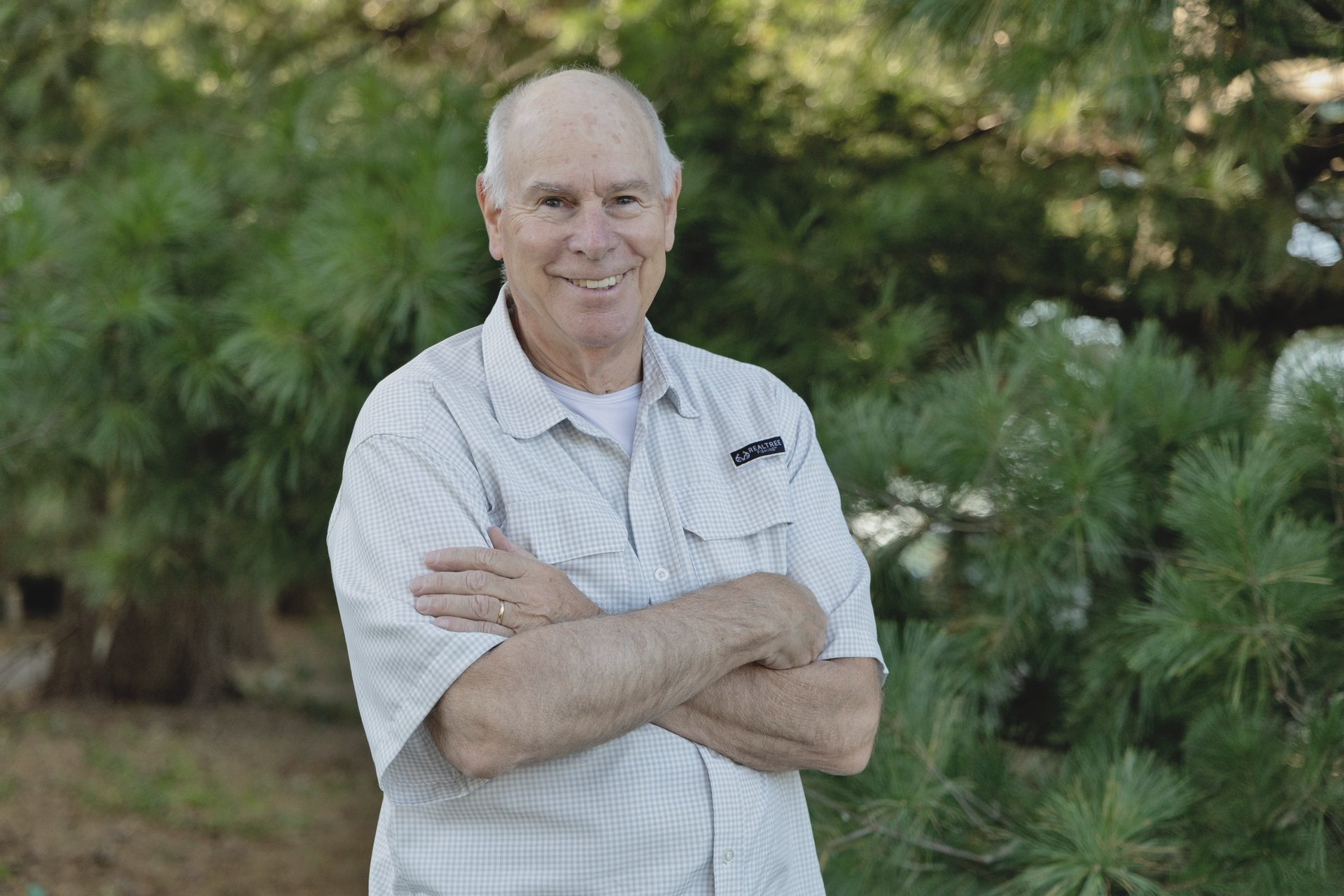 Man with crossed arms smiling in front of green foliage.