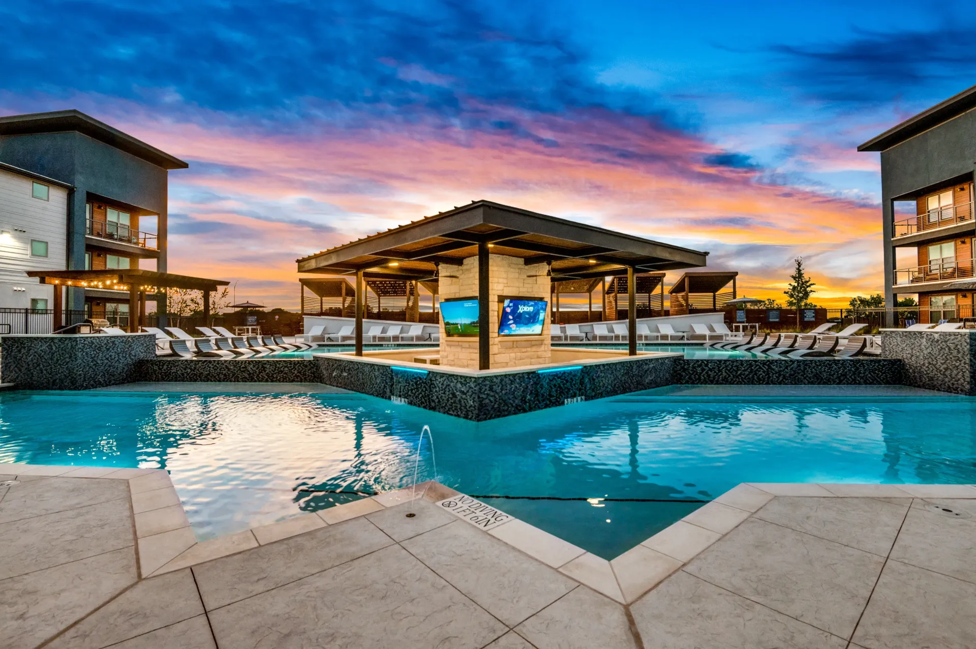 Luxury apartment pool and lounge area at sunset with fire pit and cabanas.