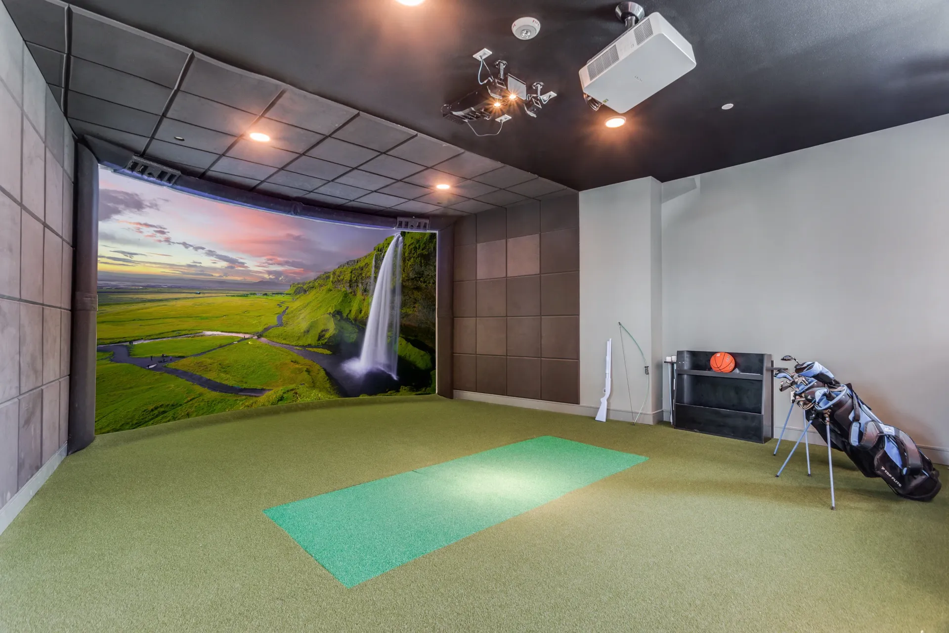 Indoor golf simulator with projection screen displaying a waterfall and landscape, golf bag, bow and arrow, and basketball.