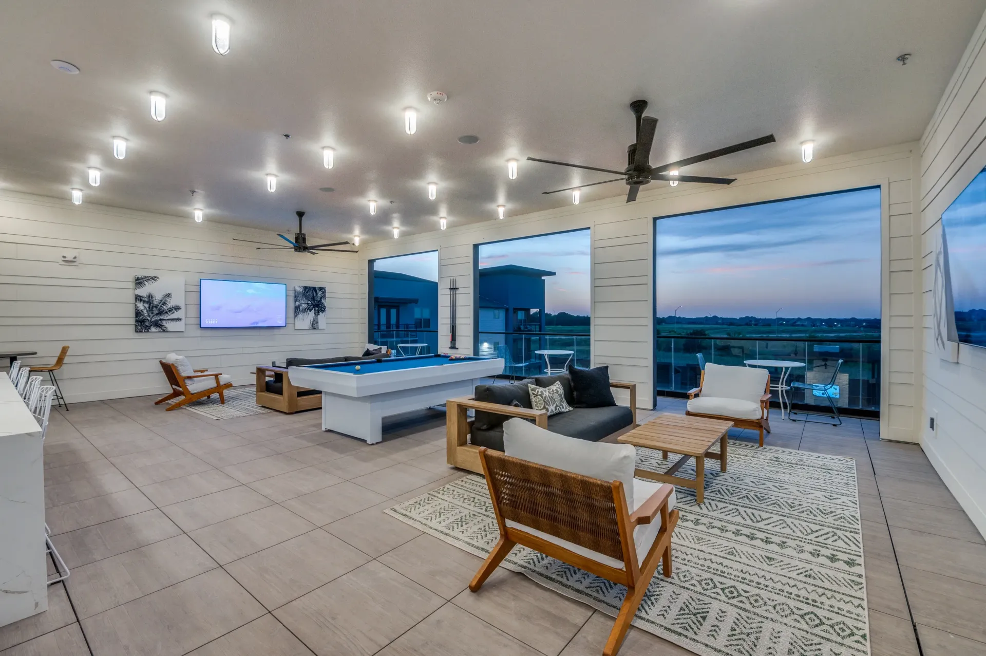 Community lounge with pool table, seating area, and large windows overlooking the outdoors at dusk.