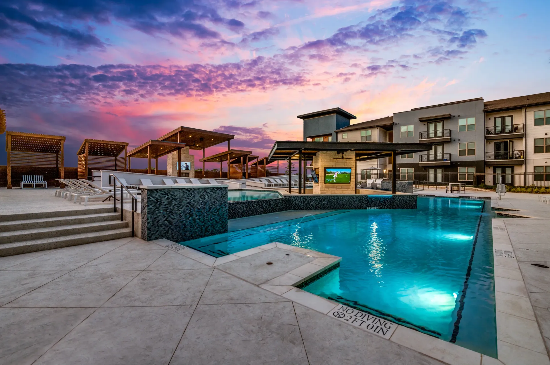 Luxury apartment pool with cabanas and lounge chairs at sunset.