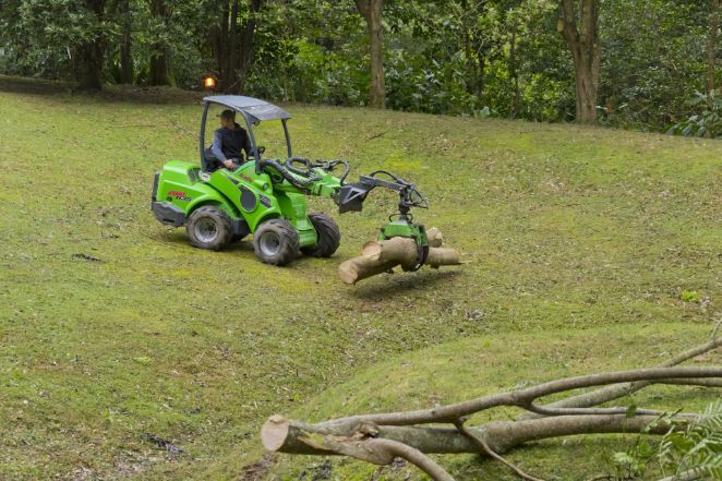 Green tractor with a log grabber lifts a log on a grassy hill — Burnett Trees in Gerringong, NSW