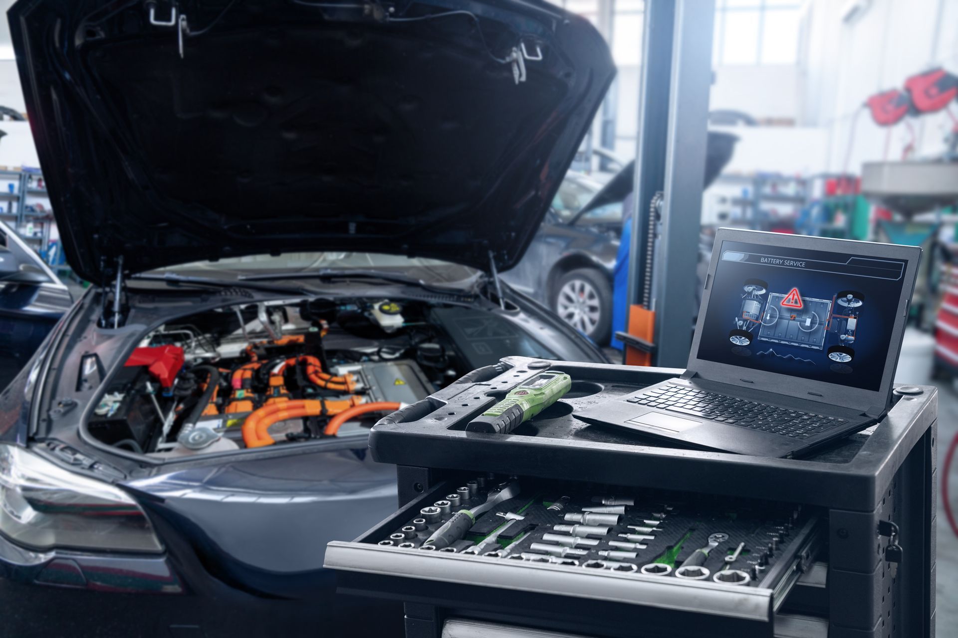 Laptop and diagnostic tools beside a car with its hood open in a garage shop.