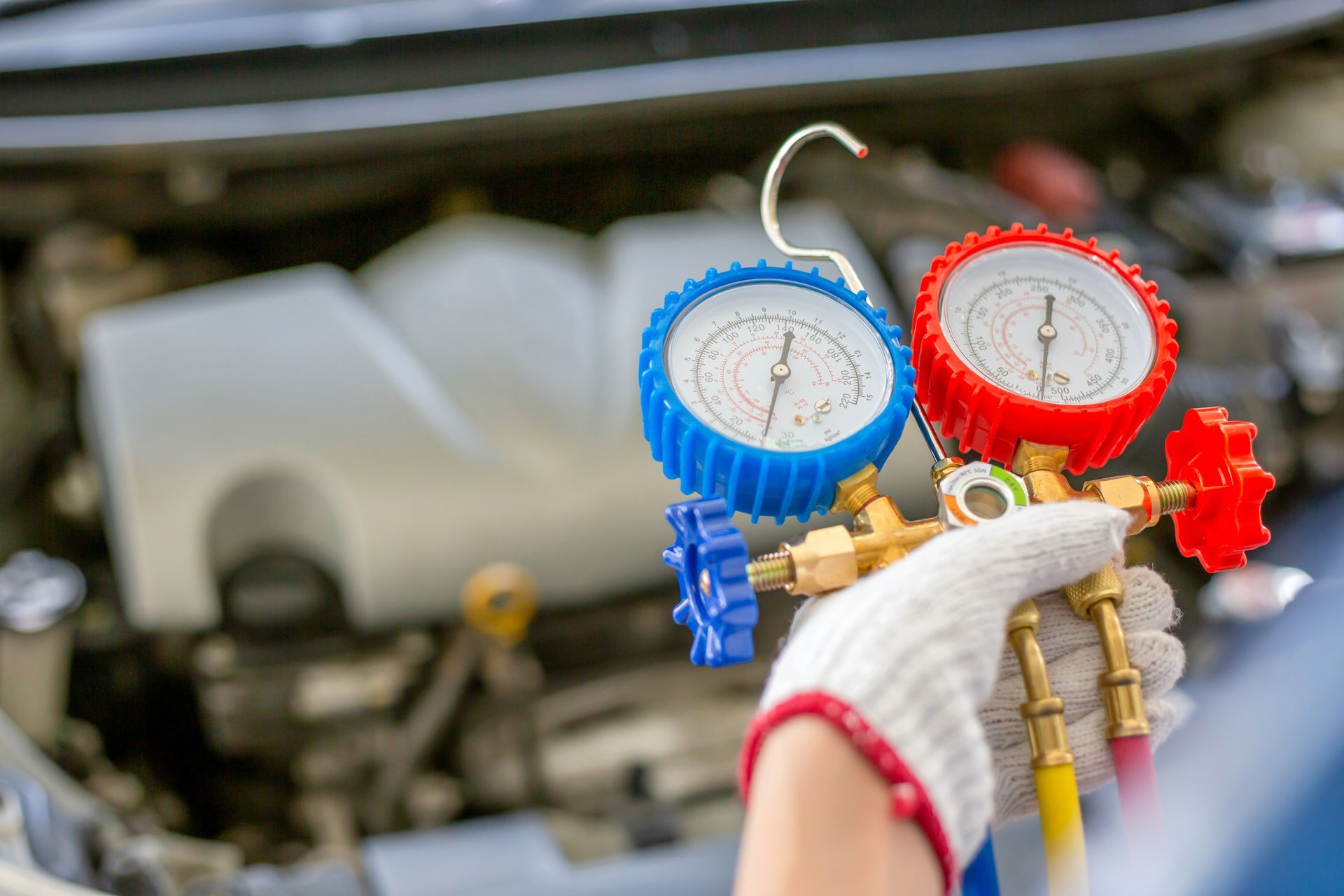 Blue and red HVAC manifold gauges connected to hoses, held by a gloved hand in a car engine bay