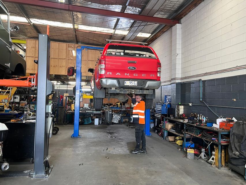 Red pickup truck raised on a lift in a mechanic’s garage, with a worker in an orange vest nearby.