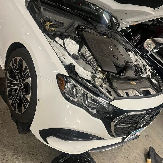 White car with severe front-end damage, hood open, exposing the engine in a garage.