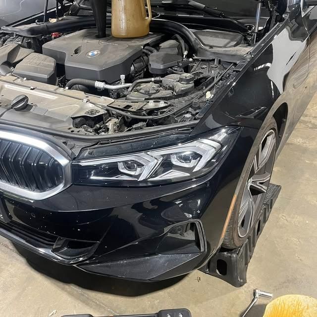 Black car with its hood open in a garage, front bumper damaged and detached on the right side.