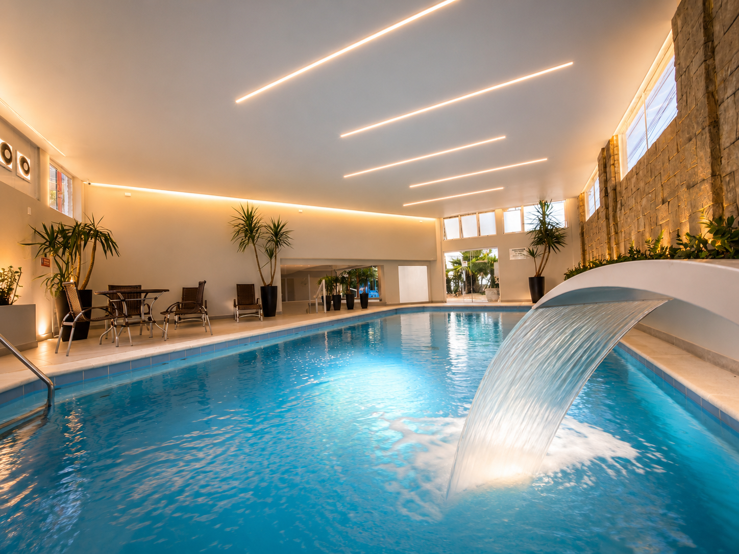 Piscina do hotel com espreguiçadeiras cor de areia, água turquesa e um prédio branco com um pátio coberto ao fundo.