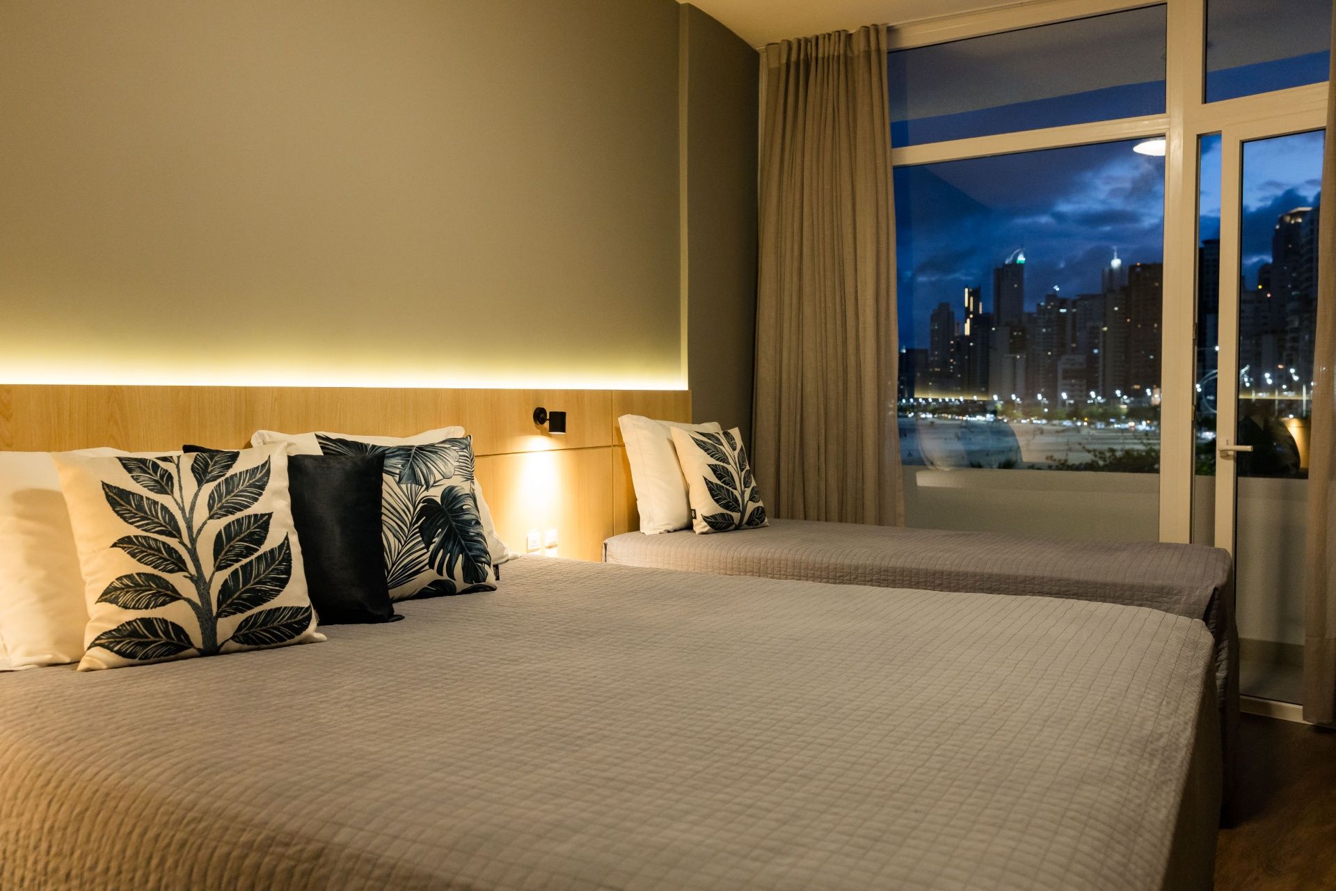 Quarto de hotel com duas camas de solteiro, decoração em tons de bege, almofadas estampadas e vista noturna da cidade pela janela.