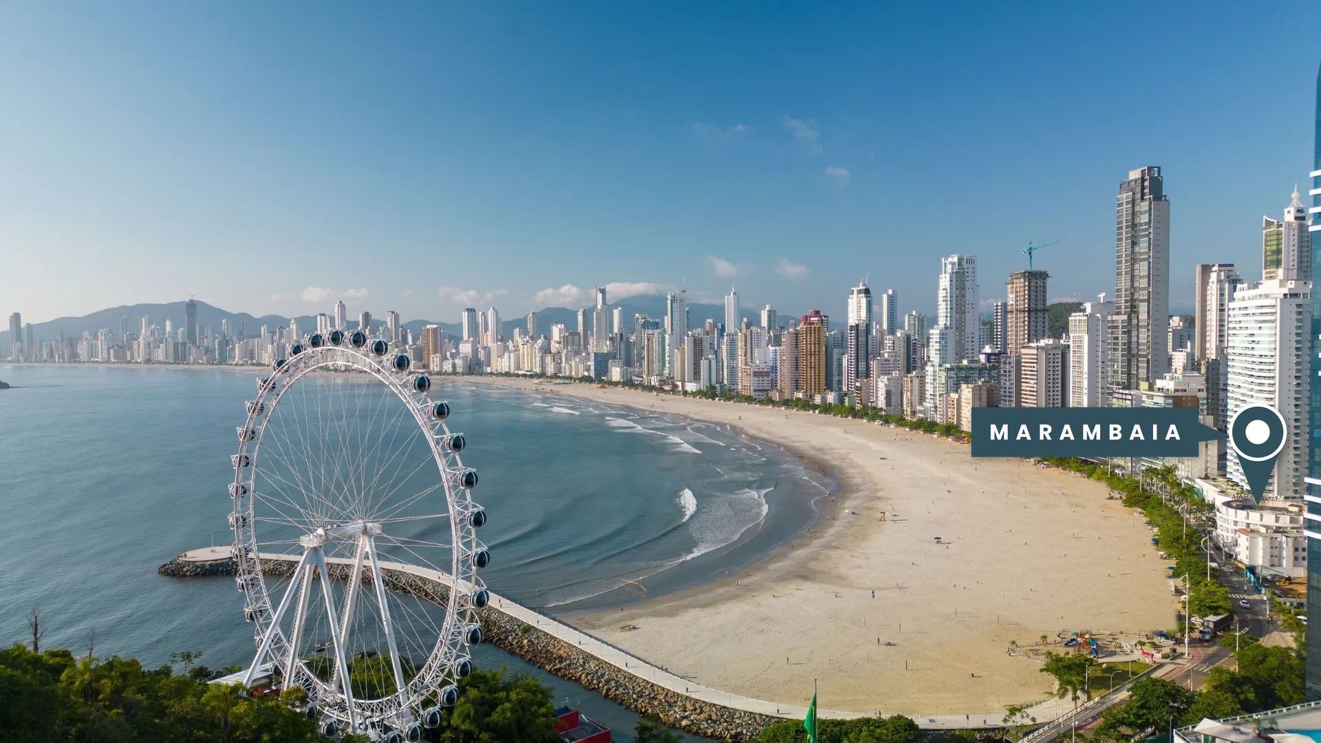 Vista aérea de uma roda-gigante e praia de areia junto a uma baía, com uma densa linha do horizonte costeiro ao fundo.