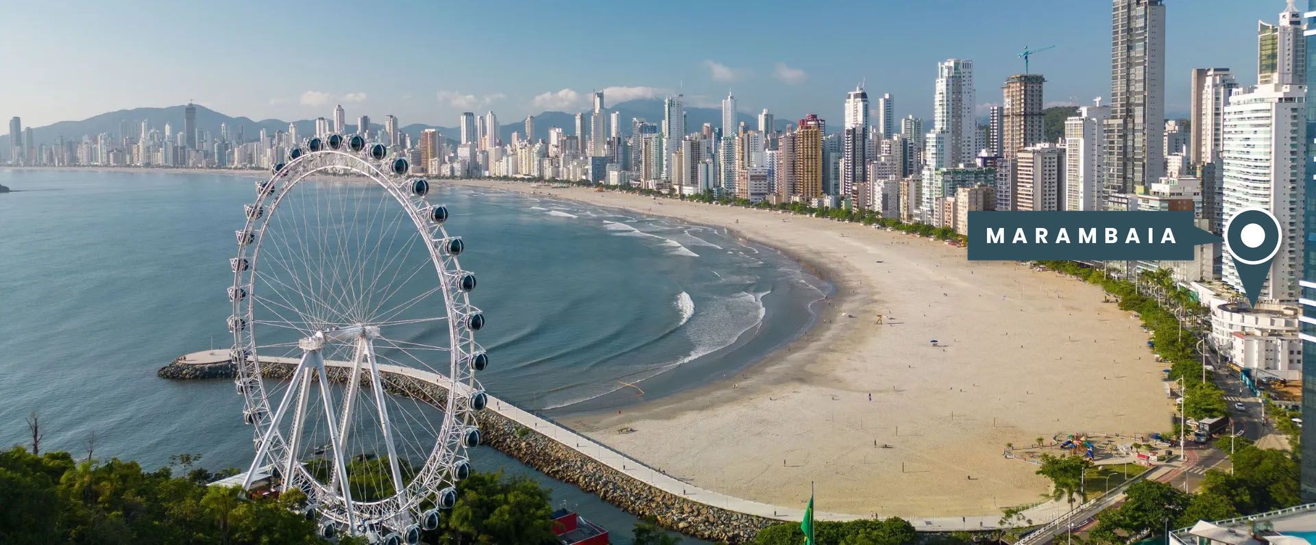 Calçadão ensolarado à beira-mar, com cúpulas brancas, árvores e altos edifícios da cidade, sob um céu azul.