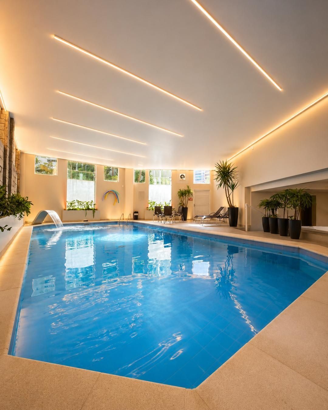 Piscina coberta com água azul-viva, luminárias elegantes no teto e plantas em vasos
