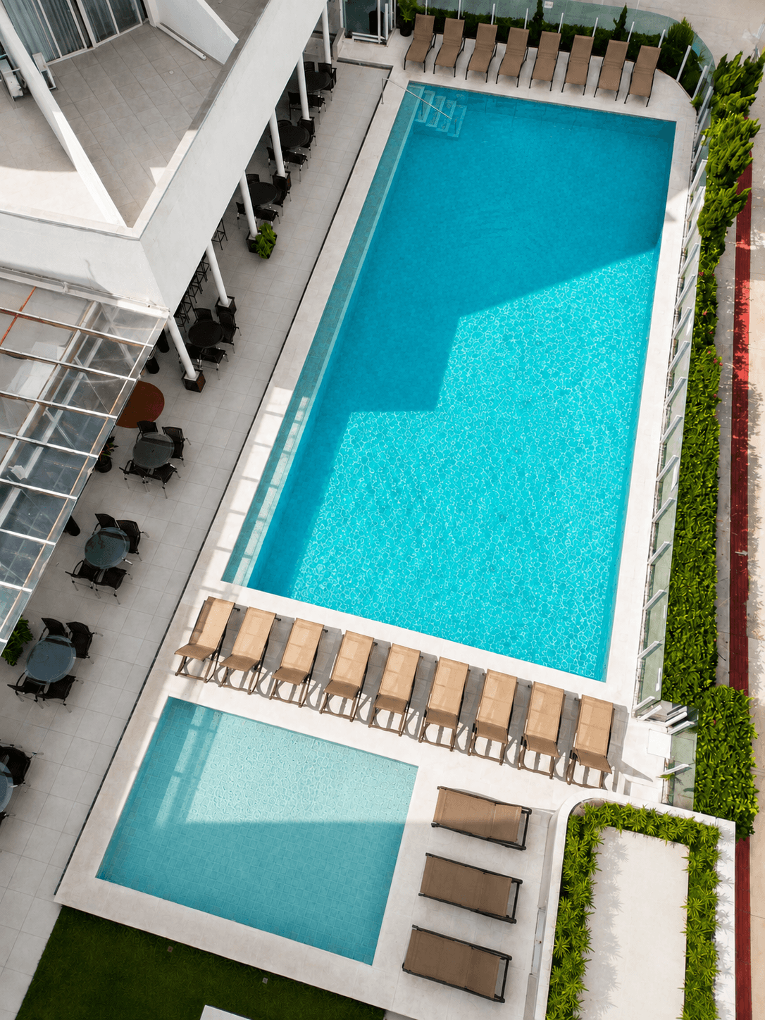 Vista aérea da área da piscina de um hotel, com uma grande piscina azul, uma piscina infantil pequena e espreguiçadeiras.