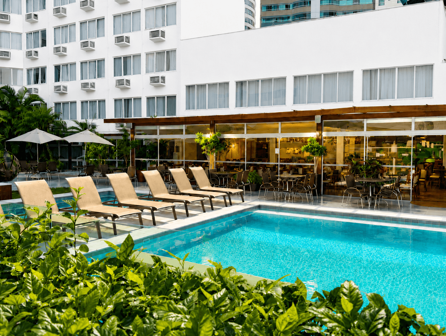 Lounge à beira da piscina de um hotel, com água azul-turquesa, cadeiras bege e vegetação exuberante diante de um edifício branco
