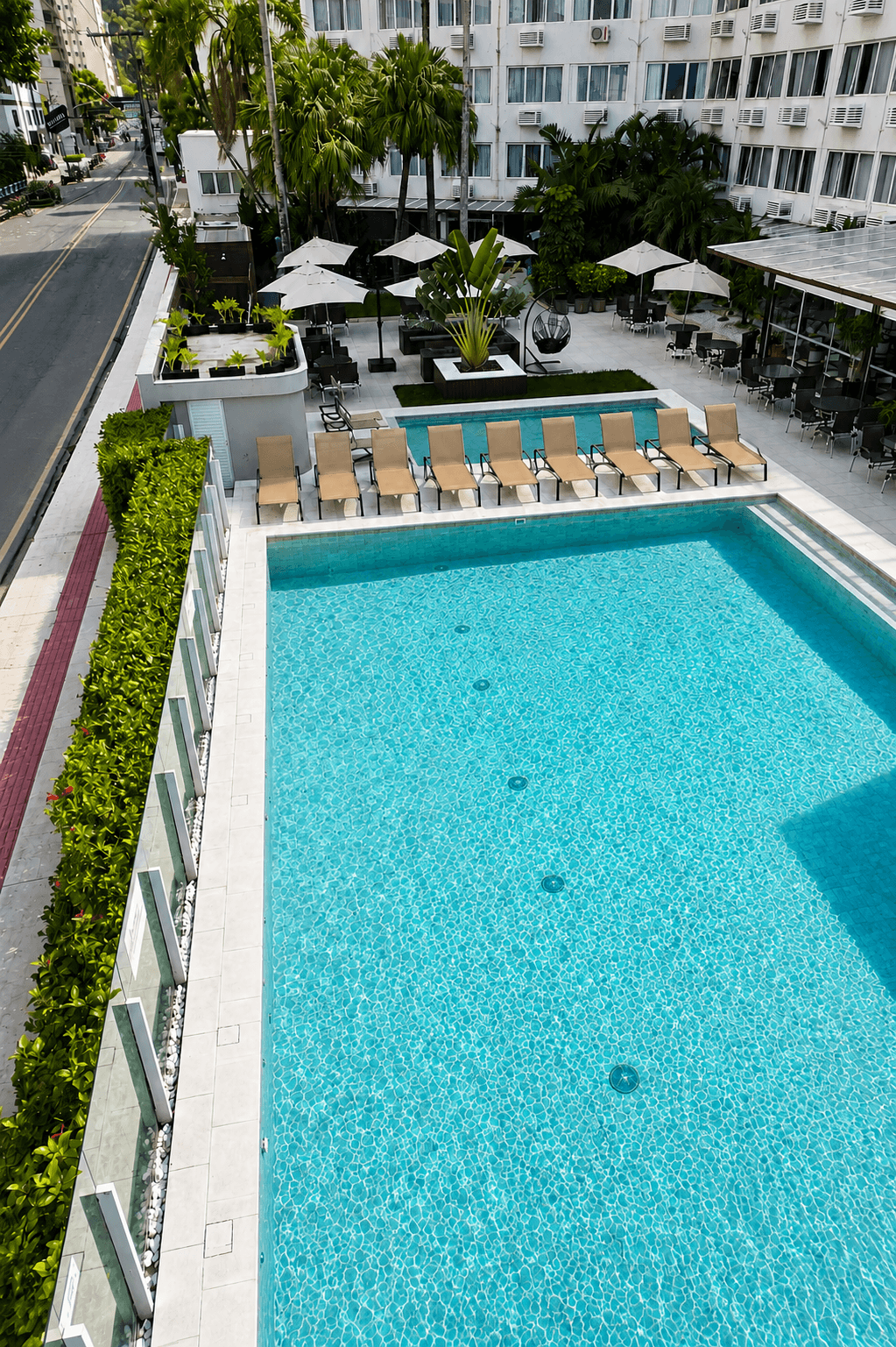 Piscina exterior de hotel com água azul-turquesa, espreguiçadeiras, guarda-sóis e vegetação ao redor, vista de cima