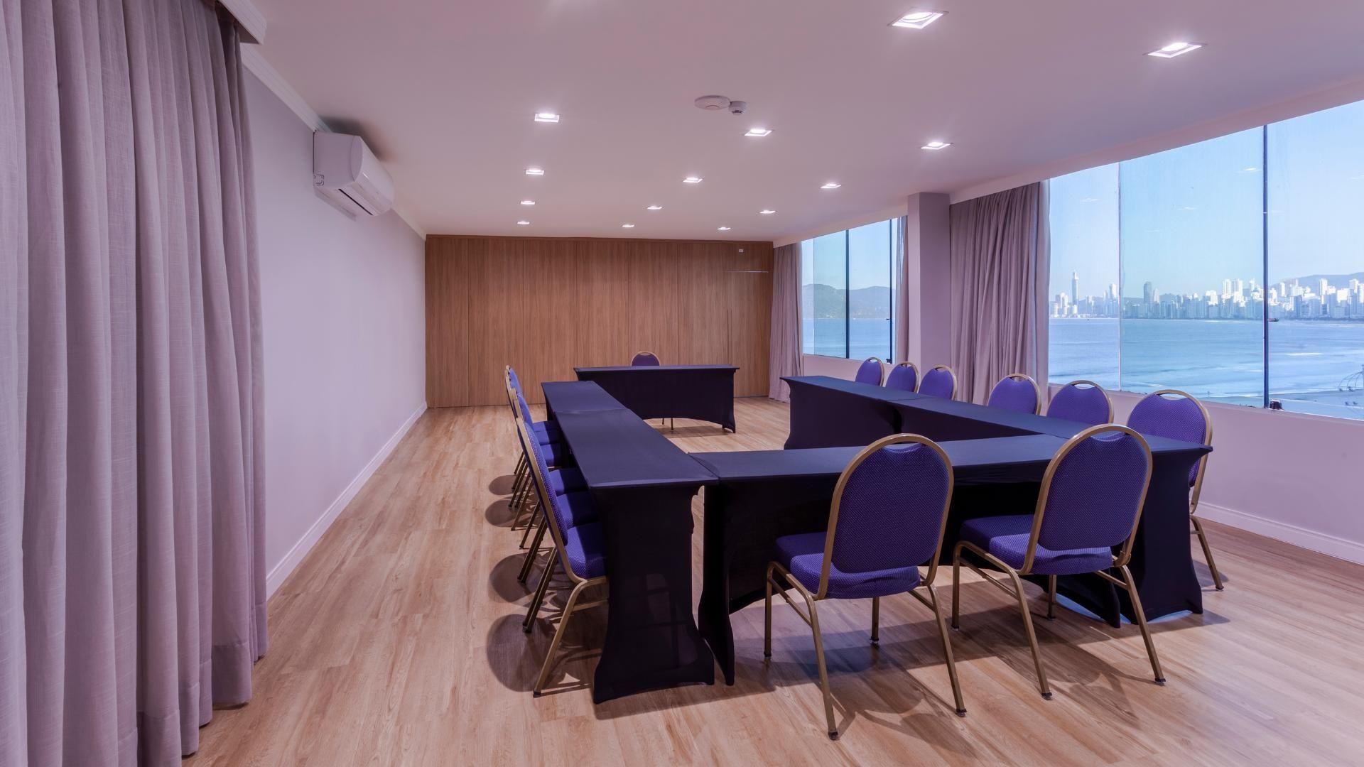 Sala de conferências vazia com fileiras de cadeiras azuis, tela de projeção e caixas de som.