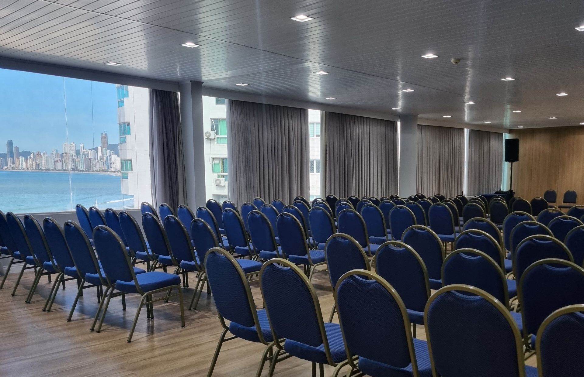 Fileiras de cadeiras azuis em uma sala de conferências iluminada, com vista para o horizonte à beira-mar