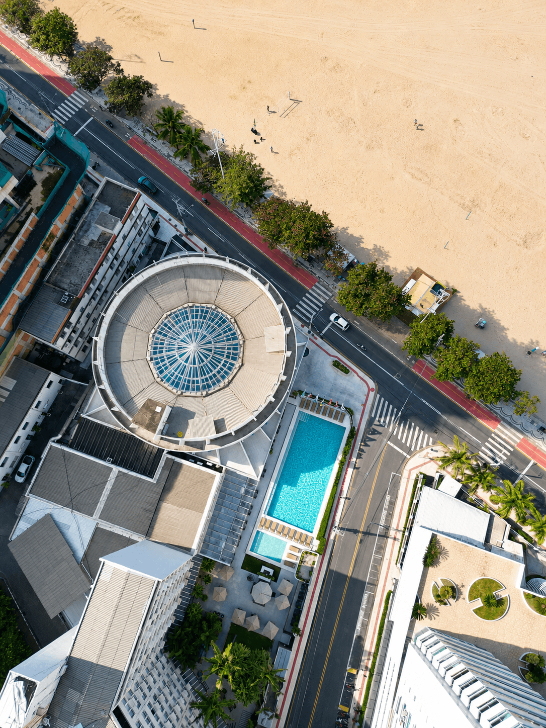 Piscina ao ar livre ao lado de um hotel branco com cadeiras e guarda-sóis turquesa em um dia ensolarado.