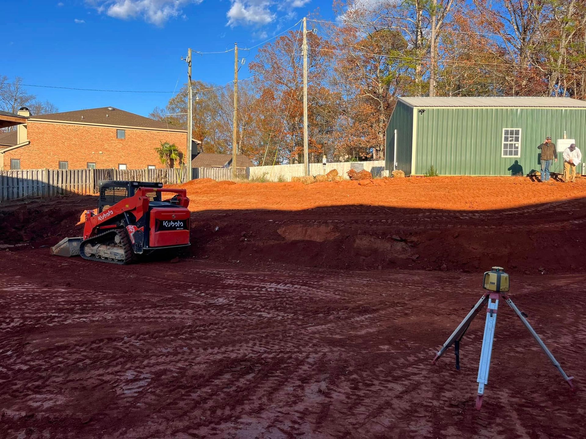 Construction site with red excavator, orange dirt, and surveying tripod under clear blue sky