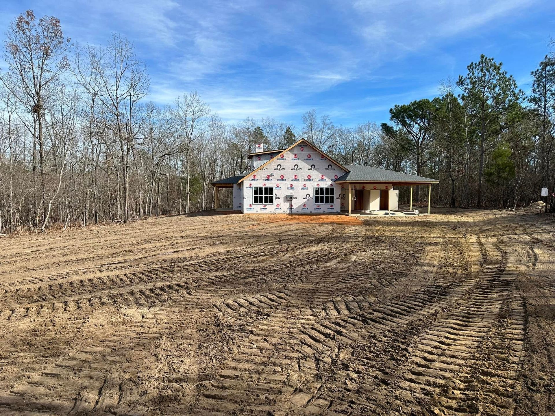 Partially built house on a cleared lot with dirt tracks and trees under a blue sky