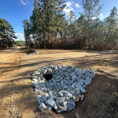 Rock-lined drainage culvert on a dirt path beside a wooded area under a blue sky