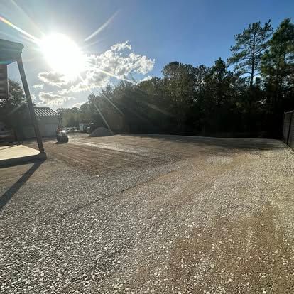 Sunlit gravel lot beside a building, with trees and a bright sky in the background
