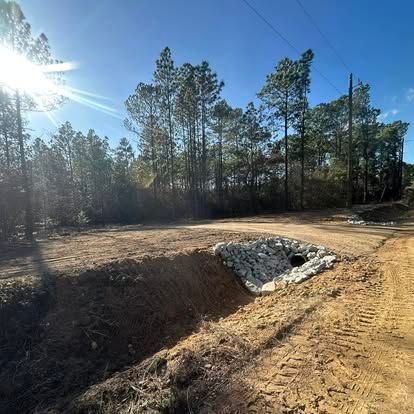 Sunlit dirt lot with a stone-lined drainage ditch beside a pine tree line