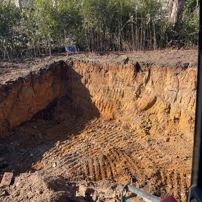 Large excavated dirt pit with steep orange-brown walls beside a wooded area