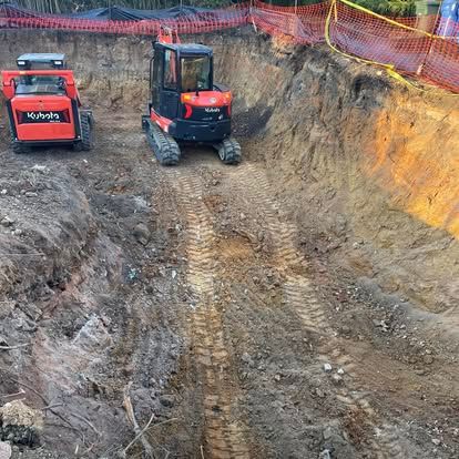 Two small excavators in a dirt excavation pit with orange safety fencing along the top edge
