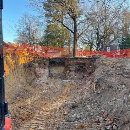 Large excavation pit at a construction site with orange safety fencing and bare trees in the background