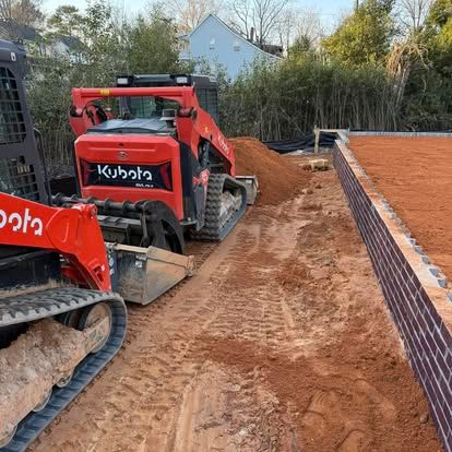 Two red Kubota skid steers grading a dirt trench beside a brick wall and fence