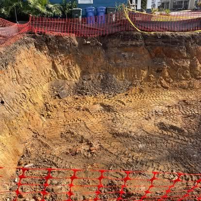 Large excavated dirt pit with orange safety fencing and construction materials along the top edge