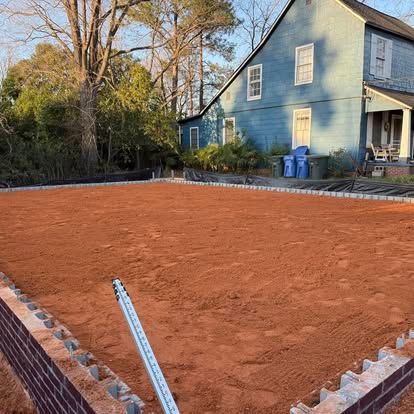 Freshly leveled red dirt lot beside a blue house, bordered by bricks and trees