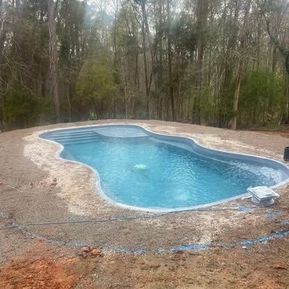 Backyard swimming pool with blue water, surrounded by gravel and trees in a wooded area