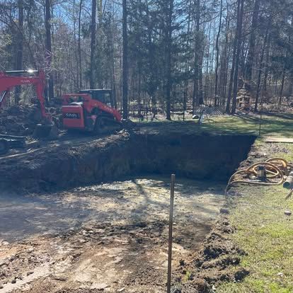 Red excavator beside a large dirt excavation pit in a wooded area