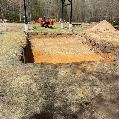 Large dug foundation pit in a grassy field, with workers and equipment in the background.