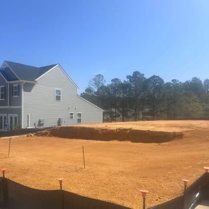 House beside a large orange dirt lot, with black silt fencing and trees under a clear blue sky