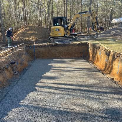 Excavation site with a yellow excavator, dirt trench, and gravel base in a wooded area.
