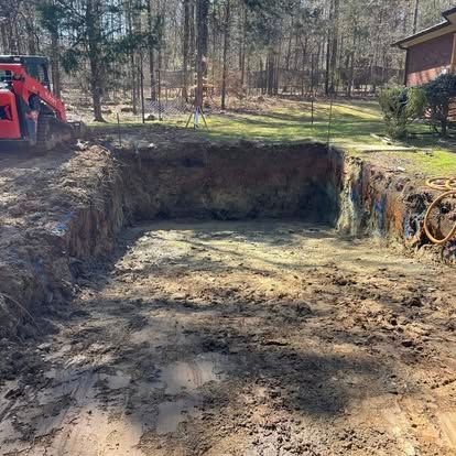 Excavated dirt pit with muddy floor, red backhoe at left, and trees and a house in the background