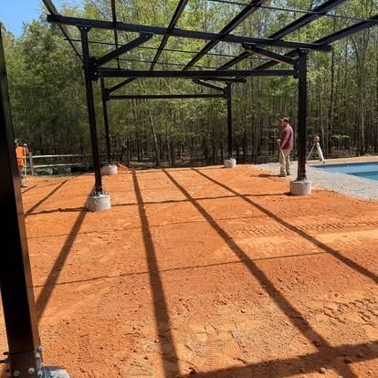 Steel frame structure on orange dirt lot, with a person standing nearby in a wooded area.