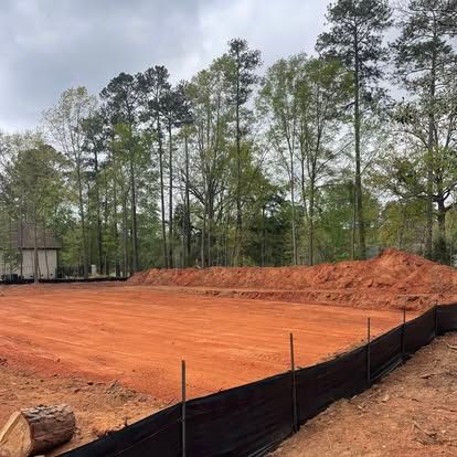 Red clay construction site beside a black silt fence and pine trees under an overcast sky