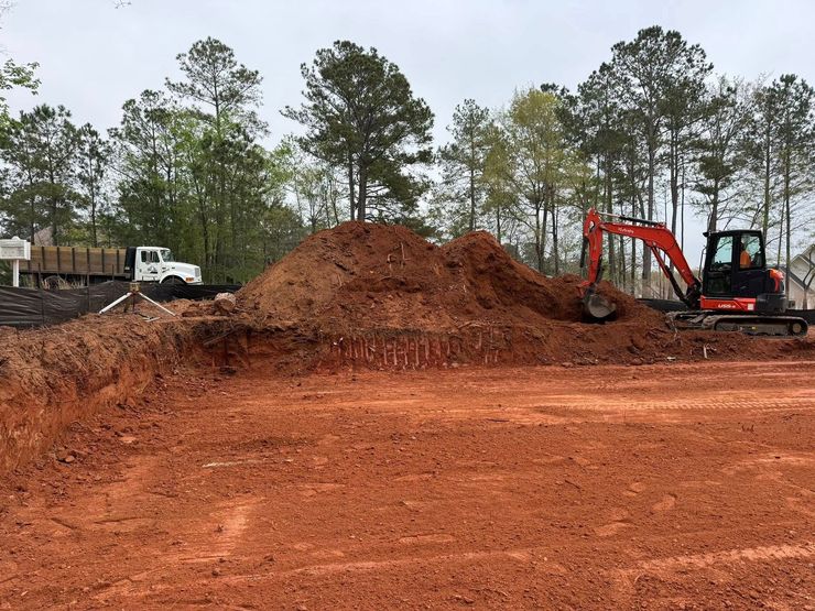 Excavator and dump truck at a red dirt construction site with a large soil mound and trees in the background