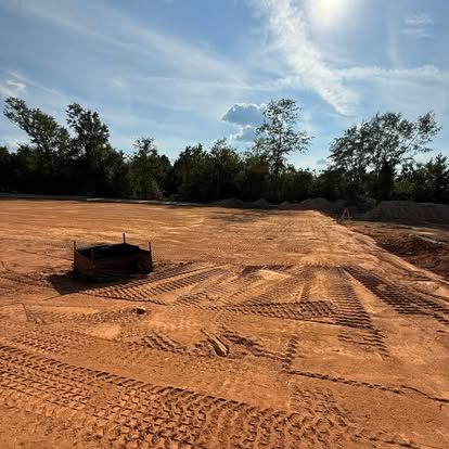 Graded dirt lot with tire tracks under a blue sky, bordered by trees and a small metal box in the foreground