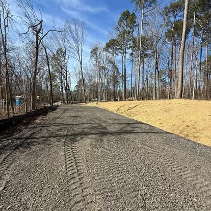 Gravel driveway through a wooded area under a clear blue sky, with bare trees and a grassy slope