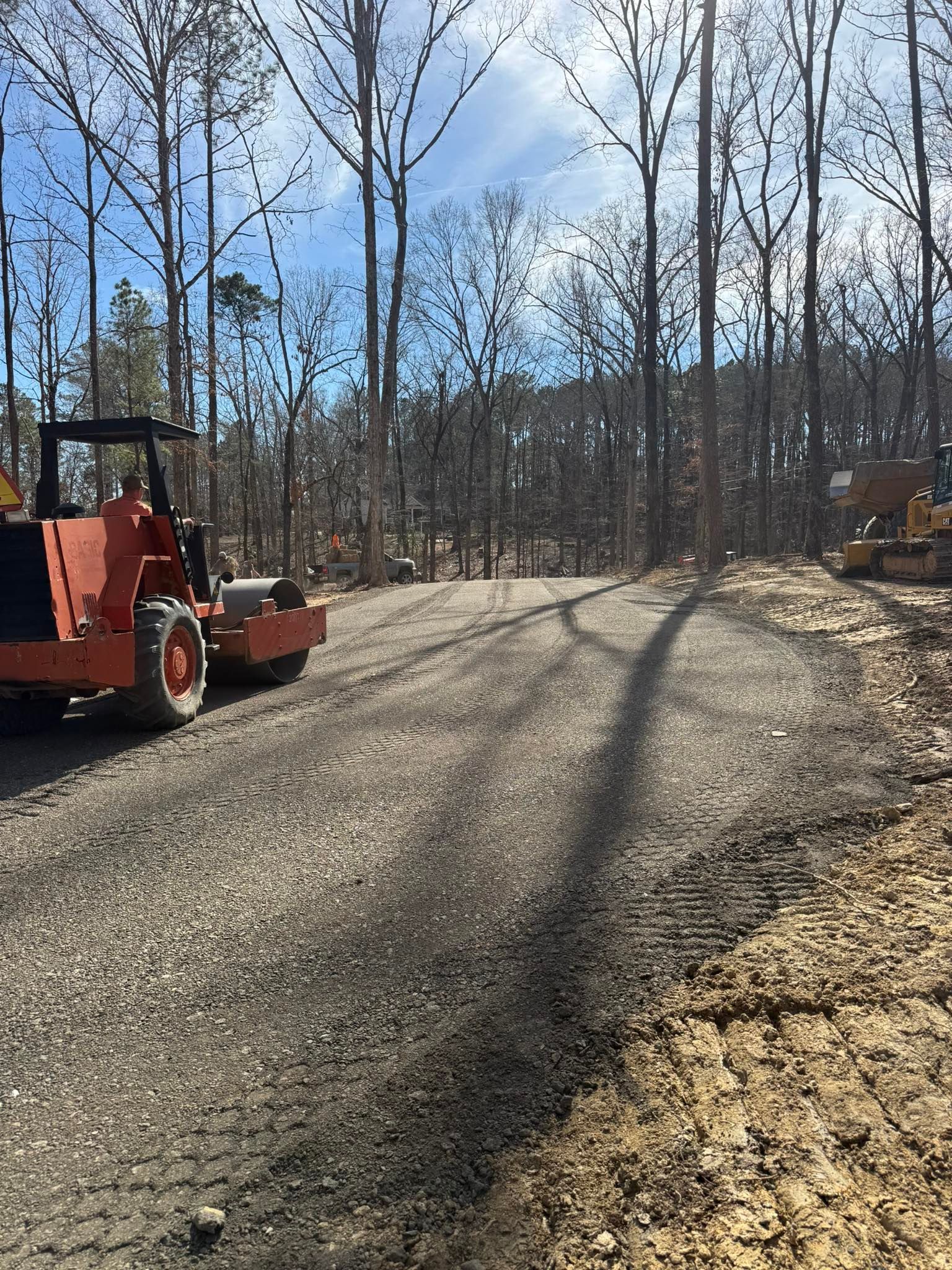 Gravel driveway in a leafless wooded area, with an orange skid steer on the left and excavator on the right.