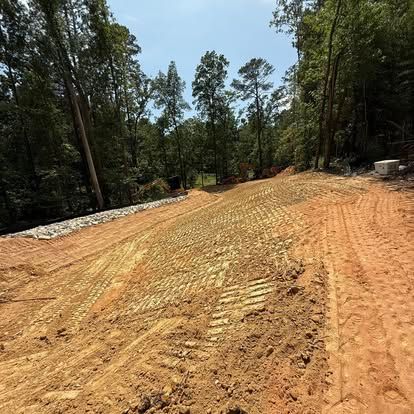 Dirt road construction site with graded orange soil, trees lining both sides, and a clear blue sky
