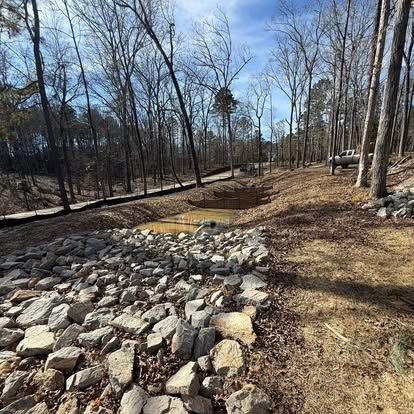 Rock-lined drainage basin in a leafless wooded area with a muddy pool and a white truck in the background