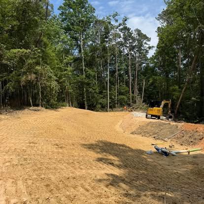 Cleared dirt lot beside trees with a yellow excavator and construction equipment in the background